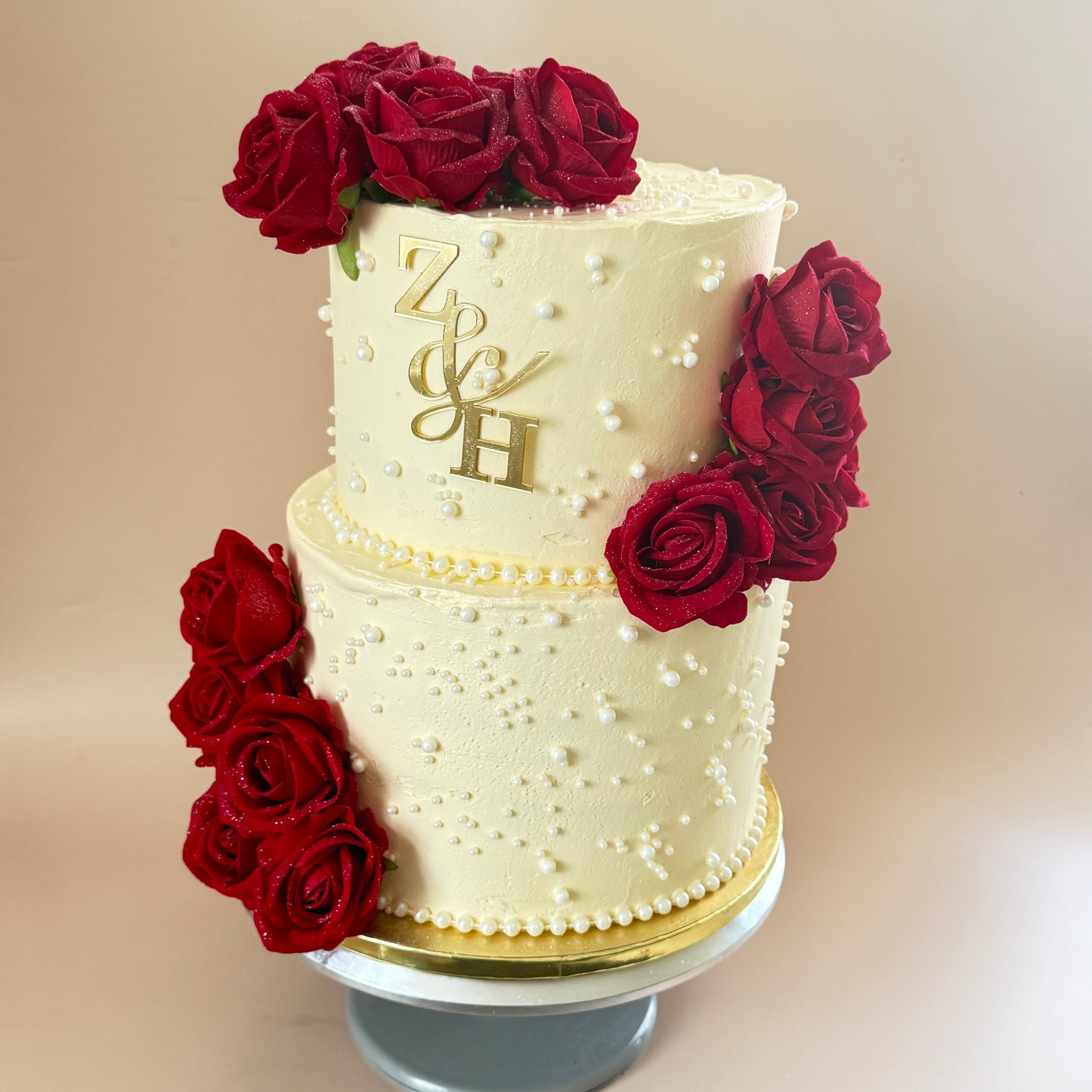 Two-tier ivory buttercream engagement cake with pearl dots, deep red roses and a gold monogram topper — custom cakes East London, Romford and Ilford