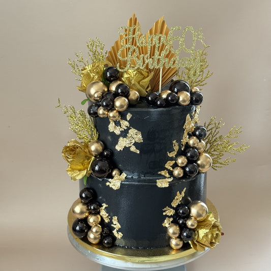 Two tier black buttercream 50th birthday cake with gold leaf, black and gold spheres, gold roses, gold paper fans and a glitter Happy 50th Birthday topper, a milestone birthday cake for Hainault IG6 by Cake Trays