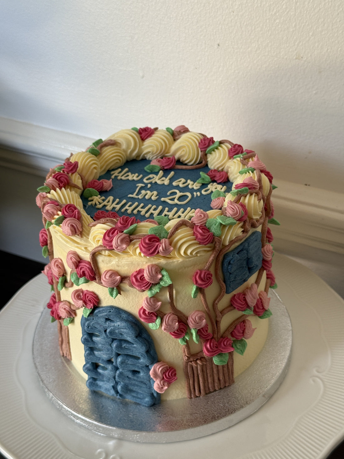 Cream buttercream birthday cake piped with pink and red roses, green leaves and a brown branch design, finished with hand piped blue plaques, made for a Forest Gate birthday in East London