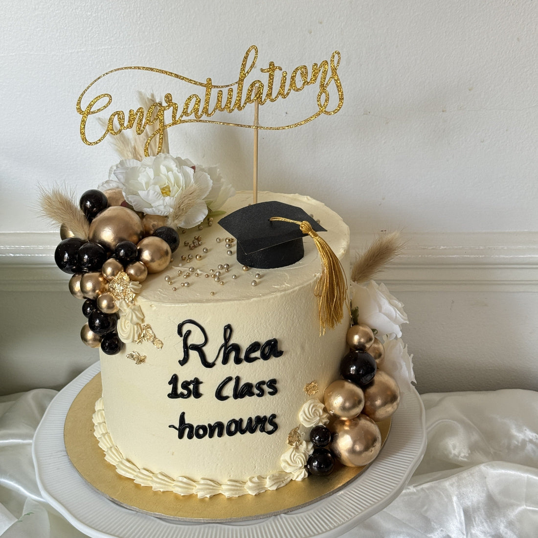 Ivory buttercream congratulations cake with a black graduation cap topper, gold and black chocolate spheres, pampas and a piped name and honours message, made for a graduate in East Ham, East London