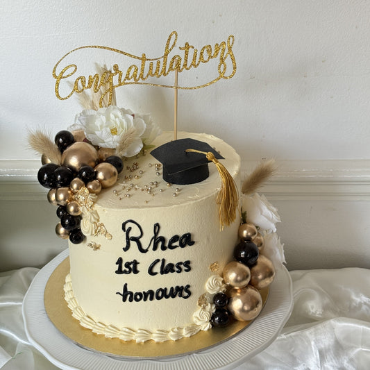 Ivory buttercream congratulations cake with a black graduation cap topper, gold and black chocolate spheres, pampas and a piped name and honours message, made for a graduate in East Ham, East London