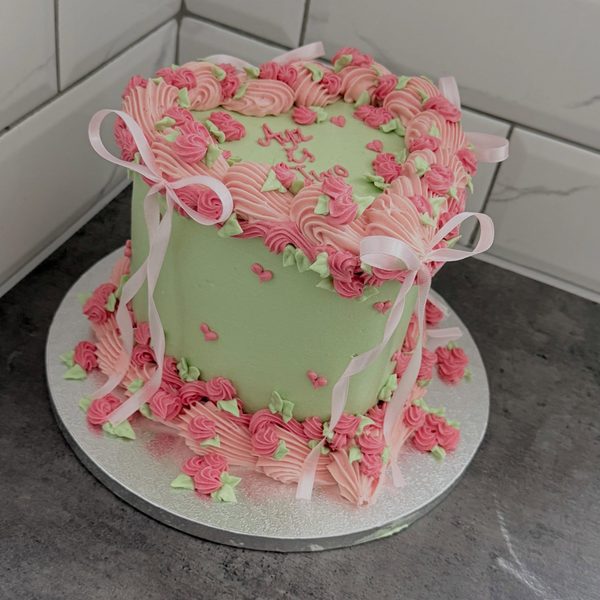 Pale green buttercream cake with pink piped rosettes, small green leaves, scattered pink hearts and pale pink satin ribbons on a silver board