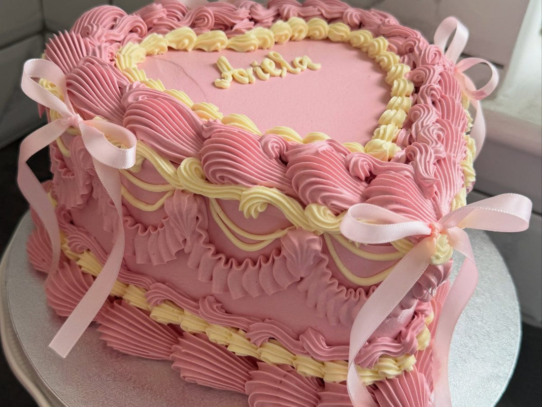 Pink heart shaped cake with pale yellow overpiped swags, pink buttercream shells, a name piped on top and pink satin bows