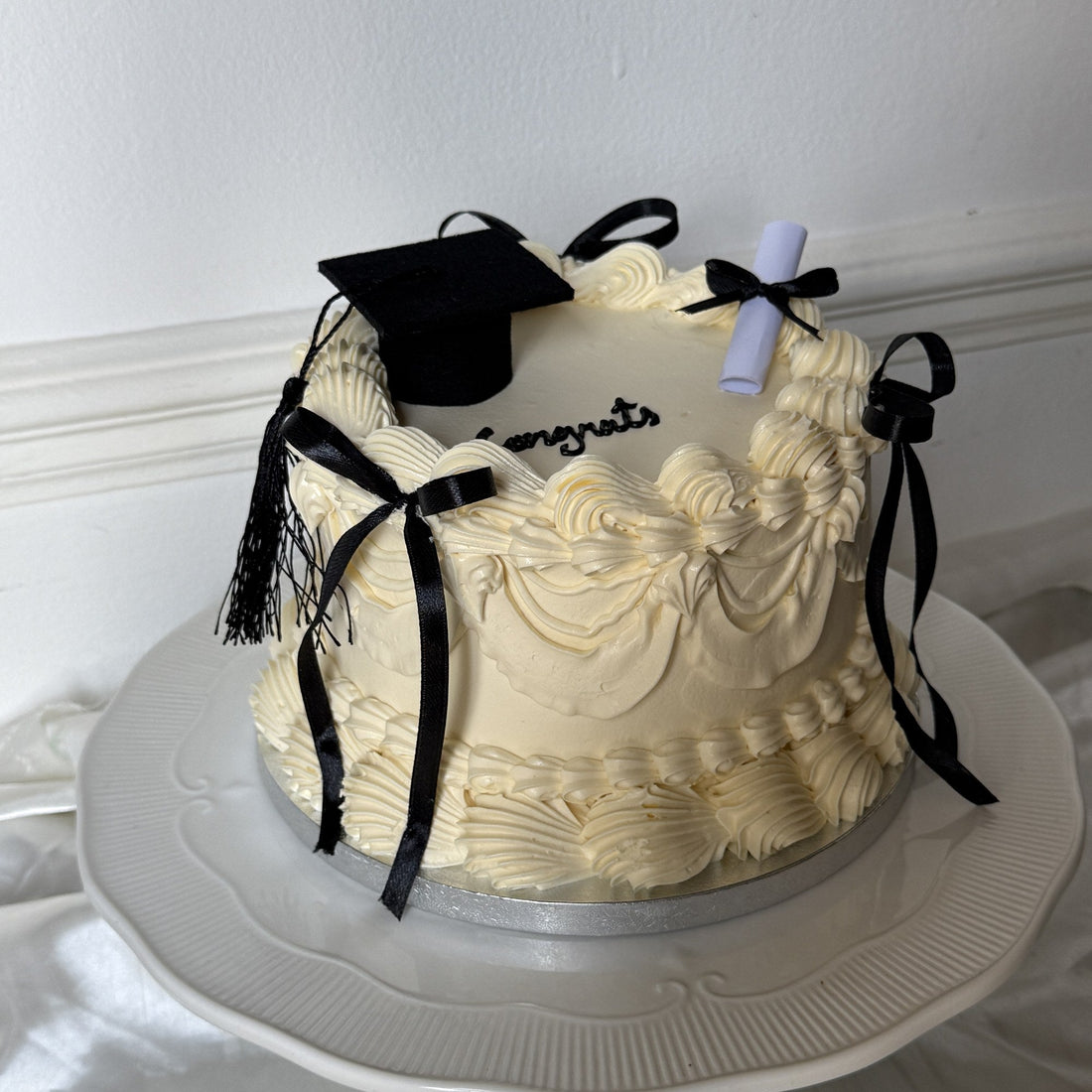 Cream buttercream graduation cake with vintage piping, black satin bows, a mortarboard topper, a paper scroll and Congrats piped on top, baked by Cake Trays in East London