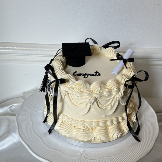 Vintage style graduation cake with ivory buttercream piping, black satin bows, a black mortarboard with tassel, a rolled scroll and Congrats piped in black, for a Stratford graduation