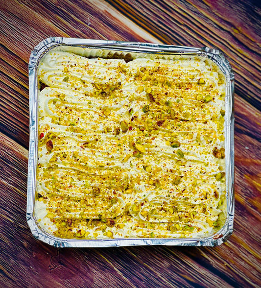 Top view of a square foil tray of pistachio milk cake, piped cream swirls covered in crushed green and gold pistachios, on a dark wooden table
