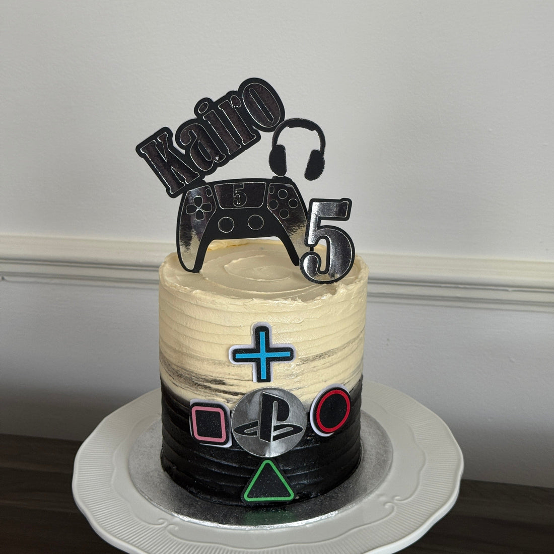 Personalised PlayStation gaming birthday cake in black and cream buttercream with a controller and headset topper and the name Kairo, made by Cake Trays in East London