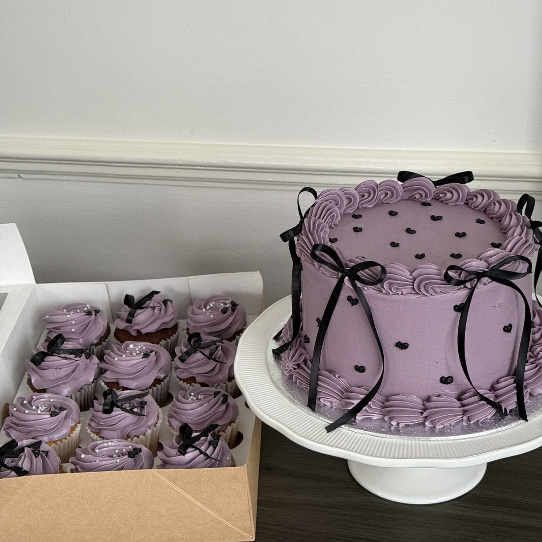 Lilac vintage buttercream bow cake with black satin bows and a matching box of bow-topped cupcakes, made by Cake Trays in East London