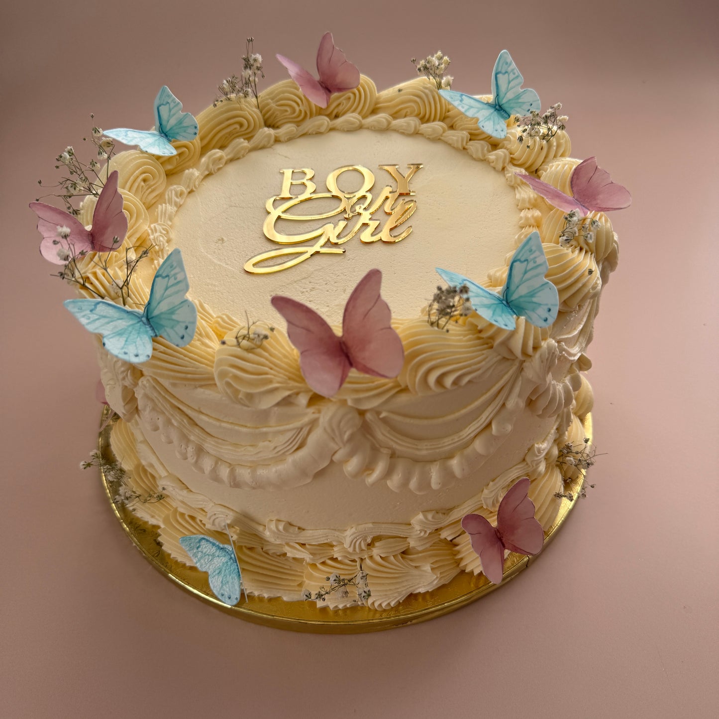 Ivory buttercream gender reveal cake with pink and blue butterflies, gypsophila and a gold Boy or Girl topper — custom cakes East London, Romford and Ilford.