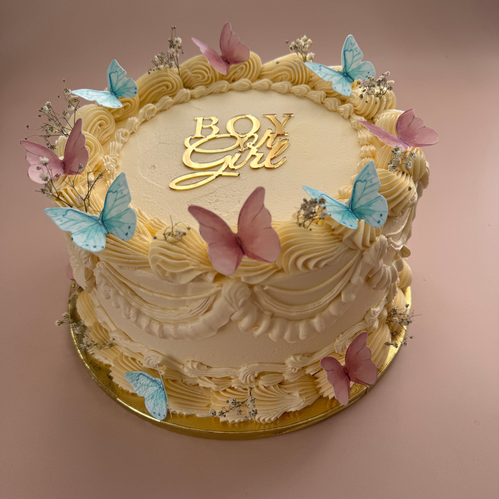Ivory buttercream gender reveal cake with pink and blue butterflies, gypsophila and a gold Boy or Girl topper — custom cakes East London, Romford and Ilford.