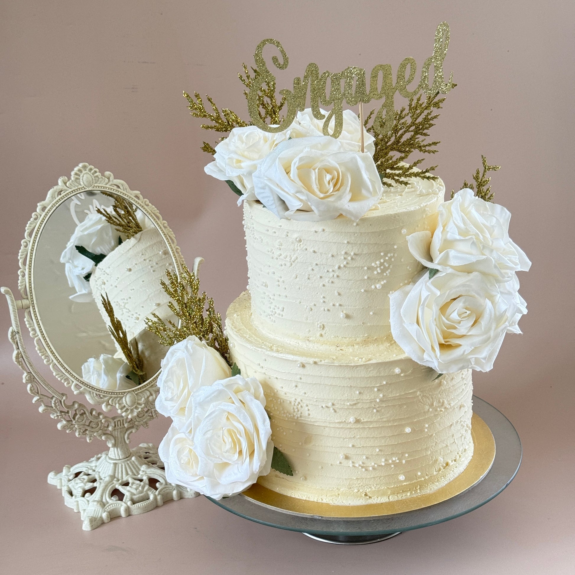 Angled view of the ivory two-tier engagement cake with white roses and gold topper — custom cakes East London, Romford and Ilford.