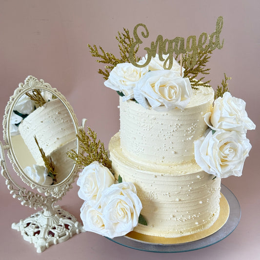 Two-tier ivory textured buttercream cake with sugar pearls, white roses and a gold Engaged topper — custom cakes East London, Romford and Ilford.
