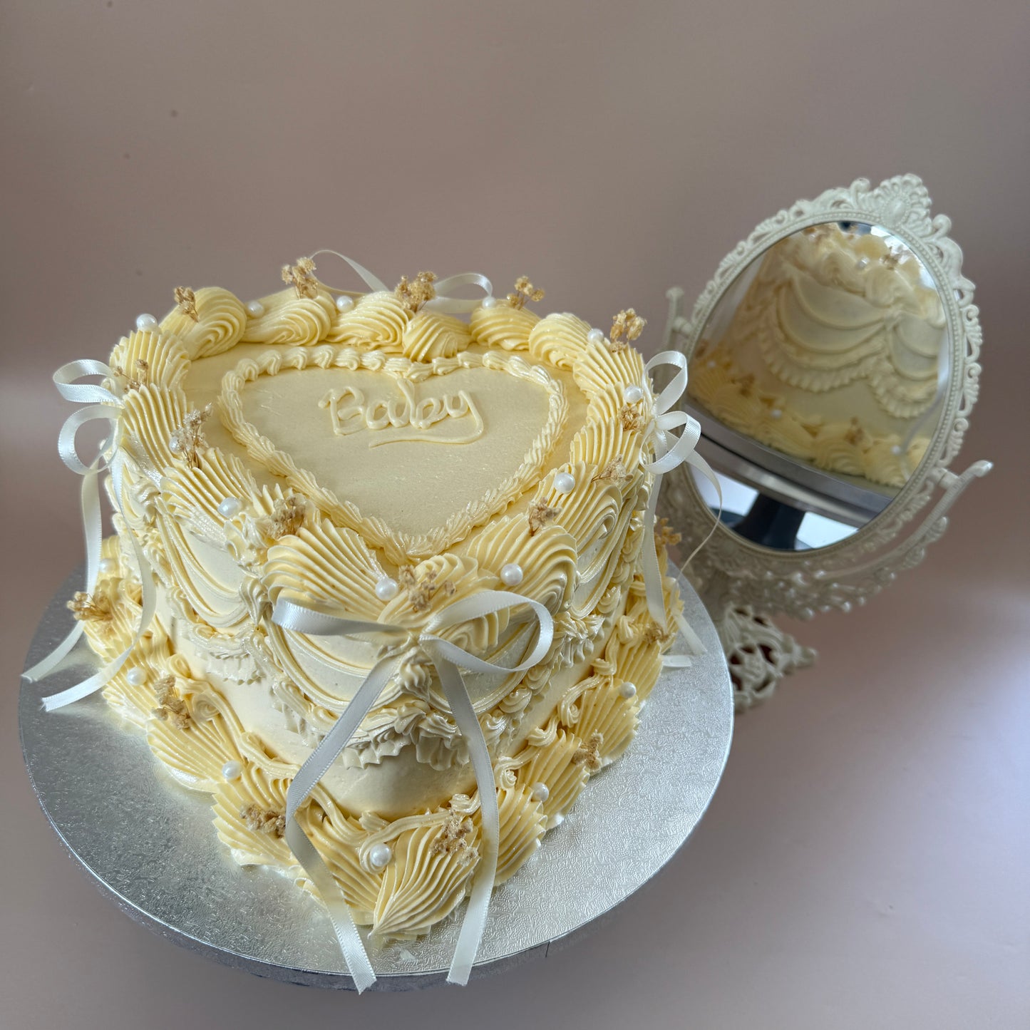 Ivory heart-shaped vintage Lambeth piped cake with pearls, ribbon bows and a 'Baby' plaque — custom cakes East London, Romford and Ilford