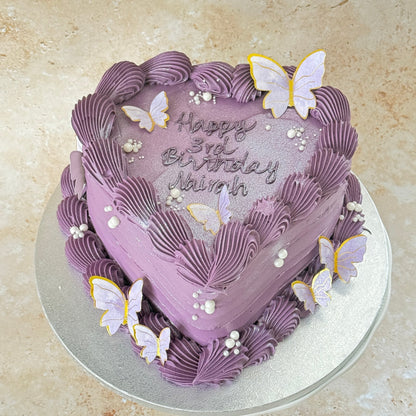 Heart-shaped lilac buttercream cake with piped shell borders, sugar pearls and gold-edged butterflies — custom cakes East London, Romford and Ilford.