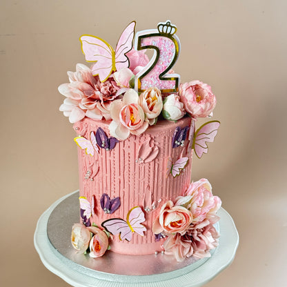 A pink frosted birthday cake decorated with sugar flowers, butterflies, and a number 2 topper, displayed on a white cake stand against a plain beige background.