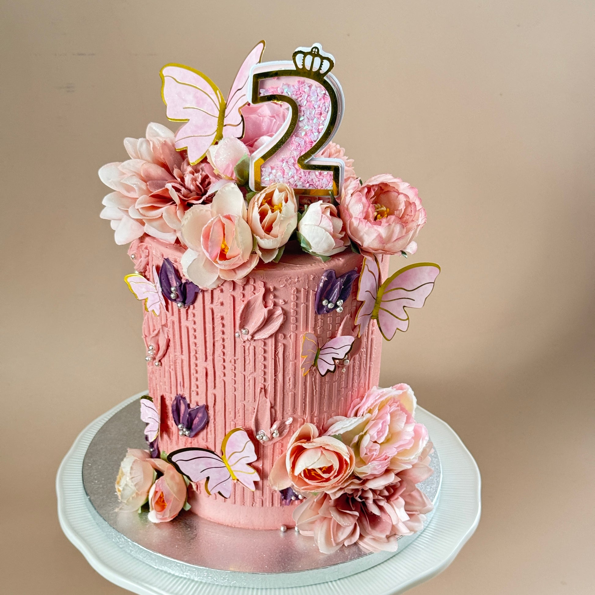 A pink frosted birthday cake decorated with sugar flowers, butterflies, and a glittery number 2 topper, displayed on a silver cake stand against a plain beige background.