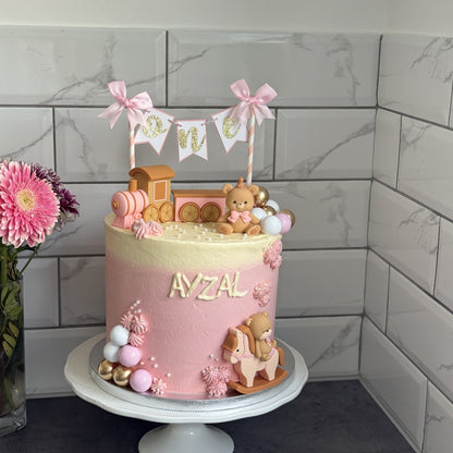 Pink teddy and train first birthday cake beside pink gerberas, custom cakes East London, Romford and Ilford.