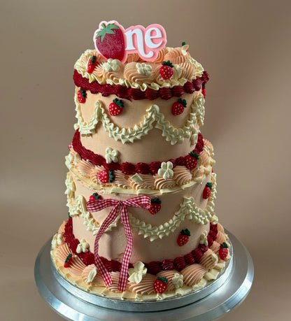 Two-tier vintage buttercream cake in peach and cream with piped ruffles, red strawberries, a gingham bow and a strawberry 'one' topper — custom cakes East London, Romford and Ilford.