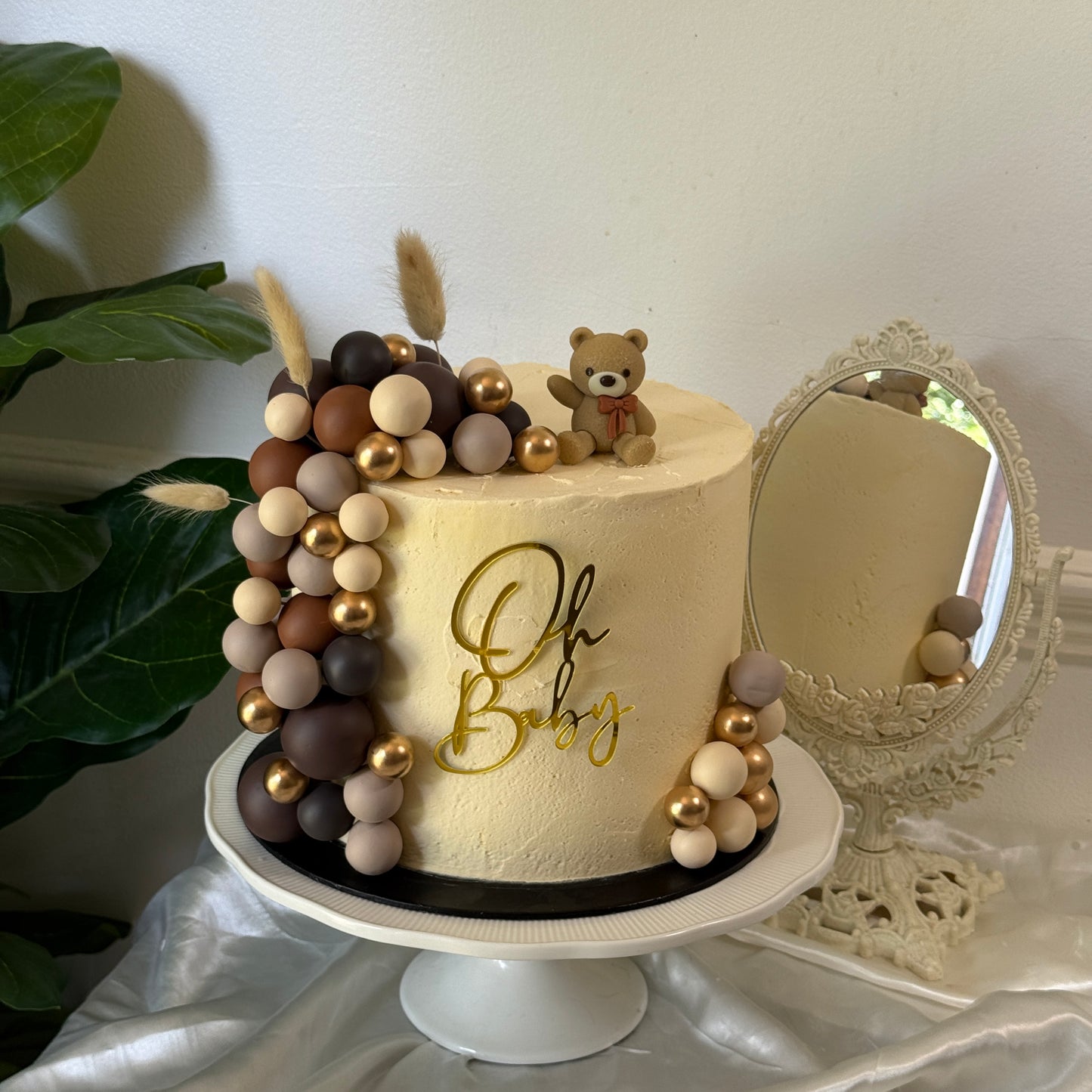 Ivory buttercream baby shower cake with brown and gold chocolate-ball garland, teddy bear topper and gold 'Oh Baby' plaque — custom cakes East London, Romford and Ilford