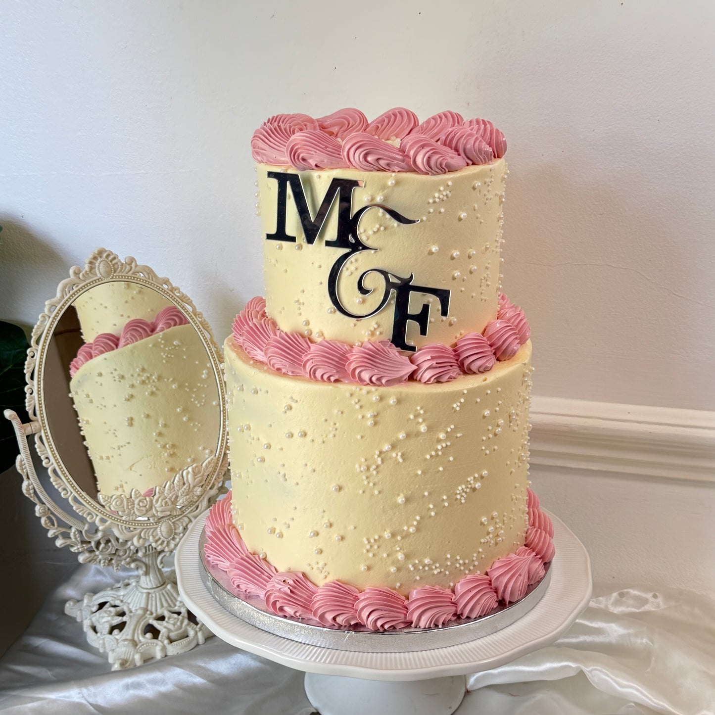 Two-tier ivory buttercream engagement cake with pink rope piping, pearl dots and a monogram topper — custom celebration cakes East London, Romford and Ilford