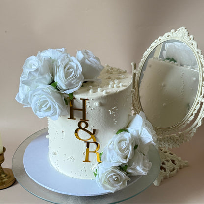 Ivory buttercream celebration cake with pearl detail, white roses and a gold initial topper — custom cakes East London, Romford and Ilford