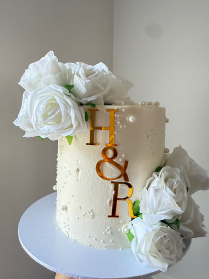 Overhead view of an ivory celebration cake with white roses and a gold initial topper — custom cakes East London, Romford and Ilford